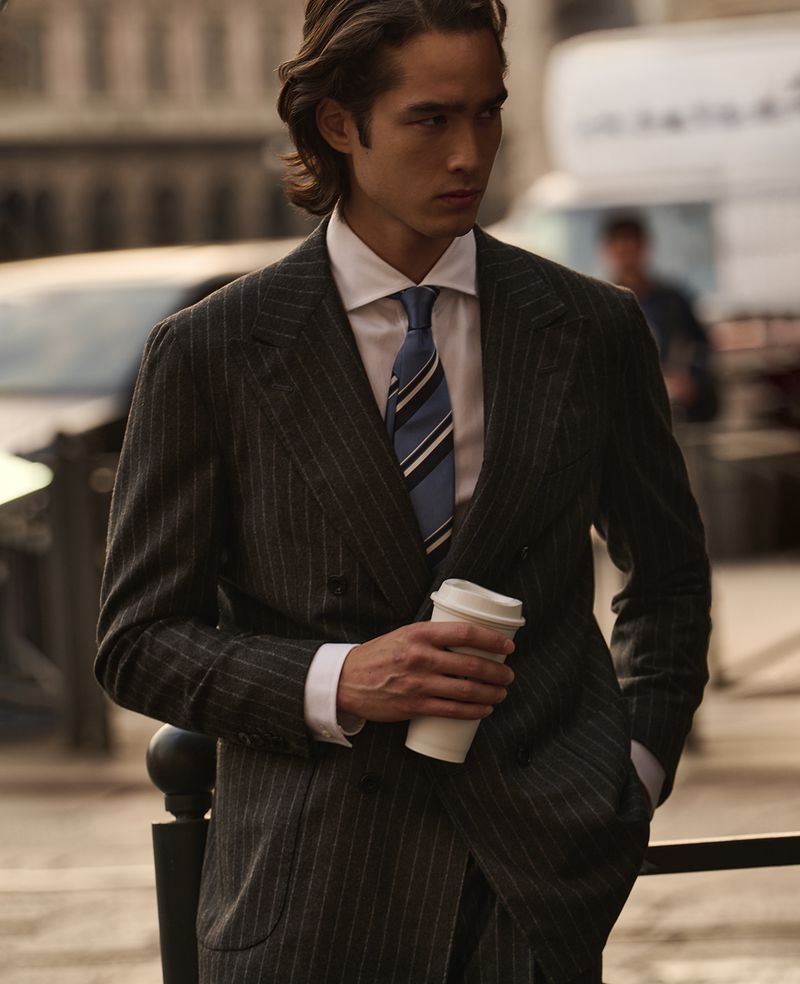 Man in a pinstripe suit stands outdoors holding a coffee cup, wearing a white shirt and striped tie. He is looking off camera.