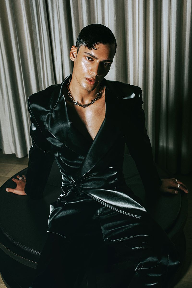 Stylish young adult male in satin black wrap-jacket and pants, wearing silver chain, posing indoors against neutral draped background.