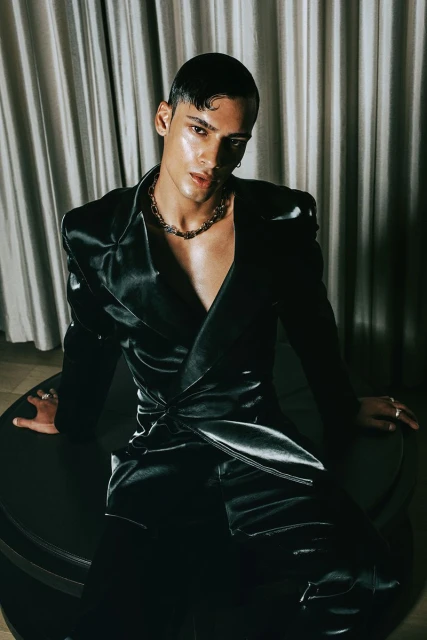Stylish young adult male in satin black wrap-jacket and pants, wearing silver chain, posing indoors against neutral draped background.
