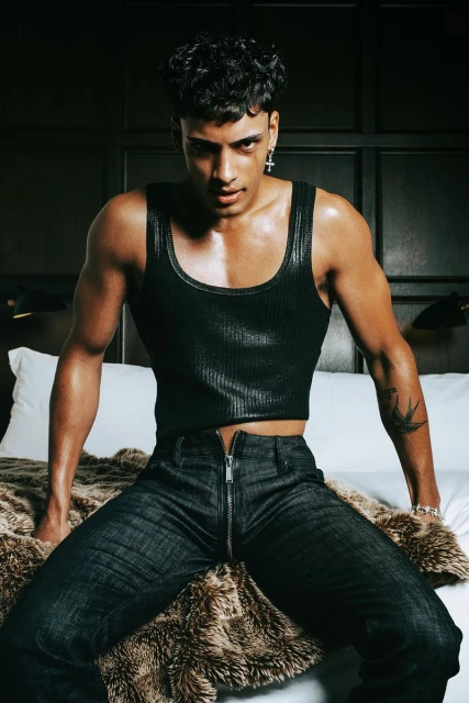 Portrait of a muscular adult male in dark tank top and jeans sitting on a bed with a faux fur blanket looking at the camera.