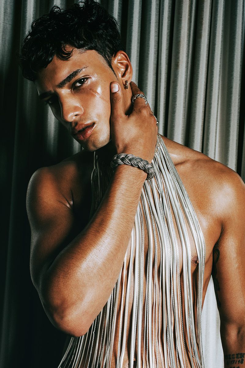 Portrait of a tan-skinned male model with wet hair, wearing a silver braided bracelet and a silver fringe top, set against draped fabric.