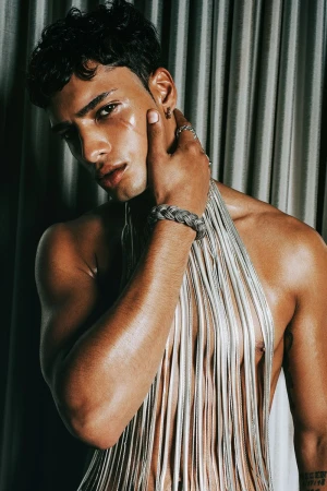 Portrait of a tan-skinned male model with wet hair, wearing a silver braided bracelet and a silver fringe top, set against draped fabric.