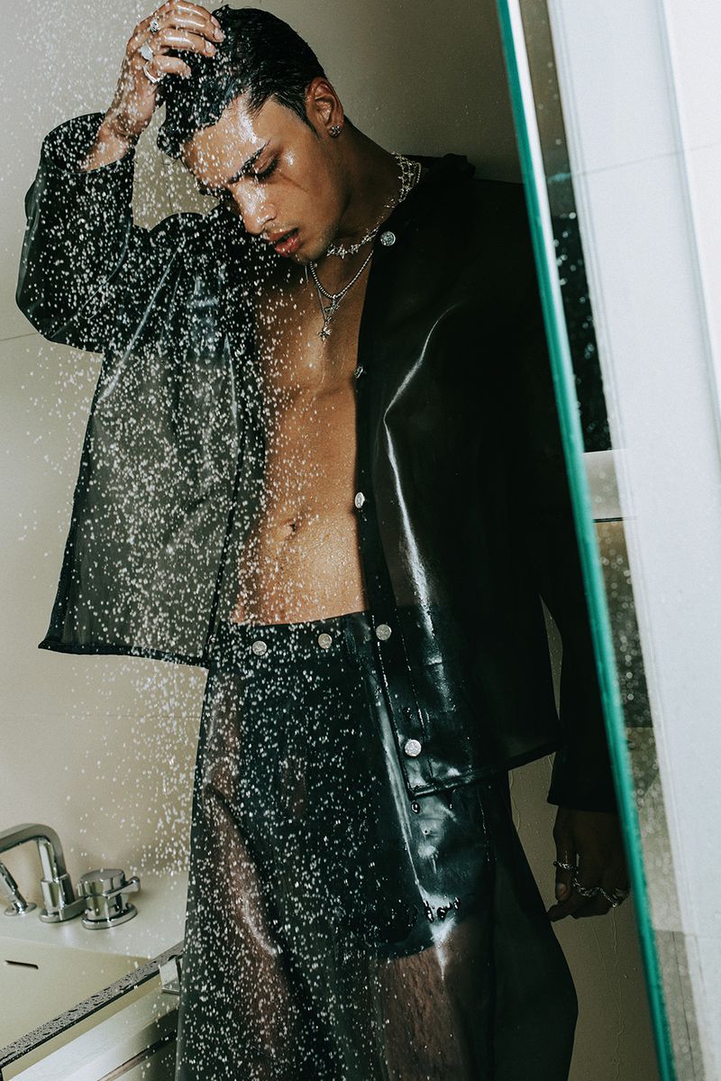 An adult male poses in a shower, wearing a sheer black plastic jacket and shorts, jewelry, with water droplets falling, for a fashion shoot.