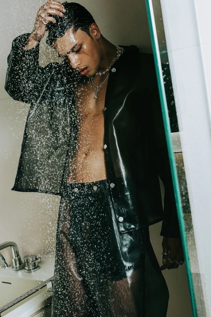 An adult male poses in a shower, wearing a sheer black plastic jacket and shorts, jewelry, with water droplets falling, for a fashion shoot.