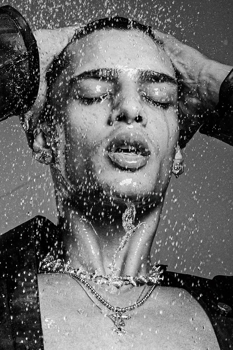 Black and white portrait of model Mase Somanlall drenched in splashing water, wearing layered necklaces and an open dark jacket.