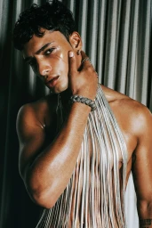 Portrait of a man with dark curly hair wearing a silver fringe halter top and jewellery against draped fabric background.
