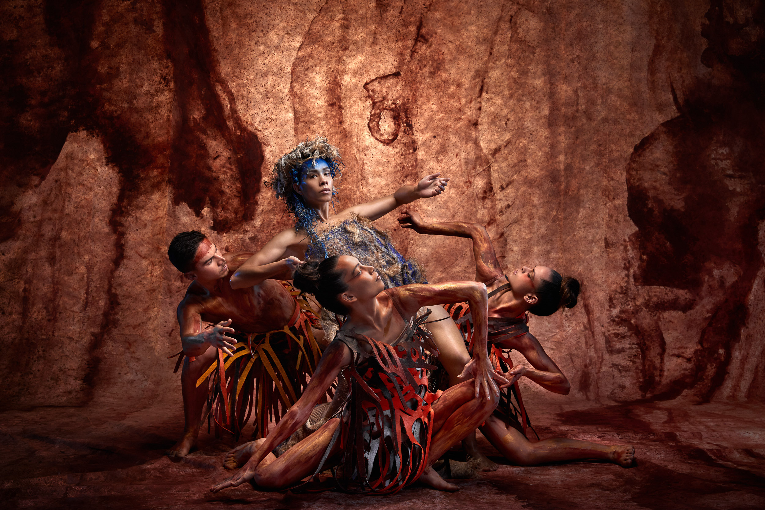 Four dancers in artistic costumes pose against a textured brown backdrop in a studio setting, showcasing expressive movements and earthy tones.