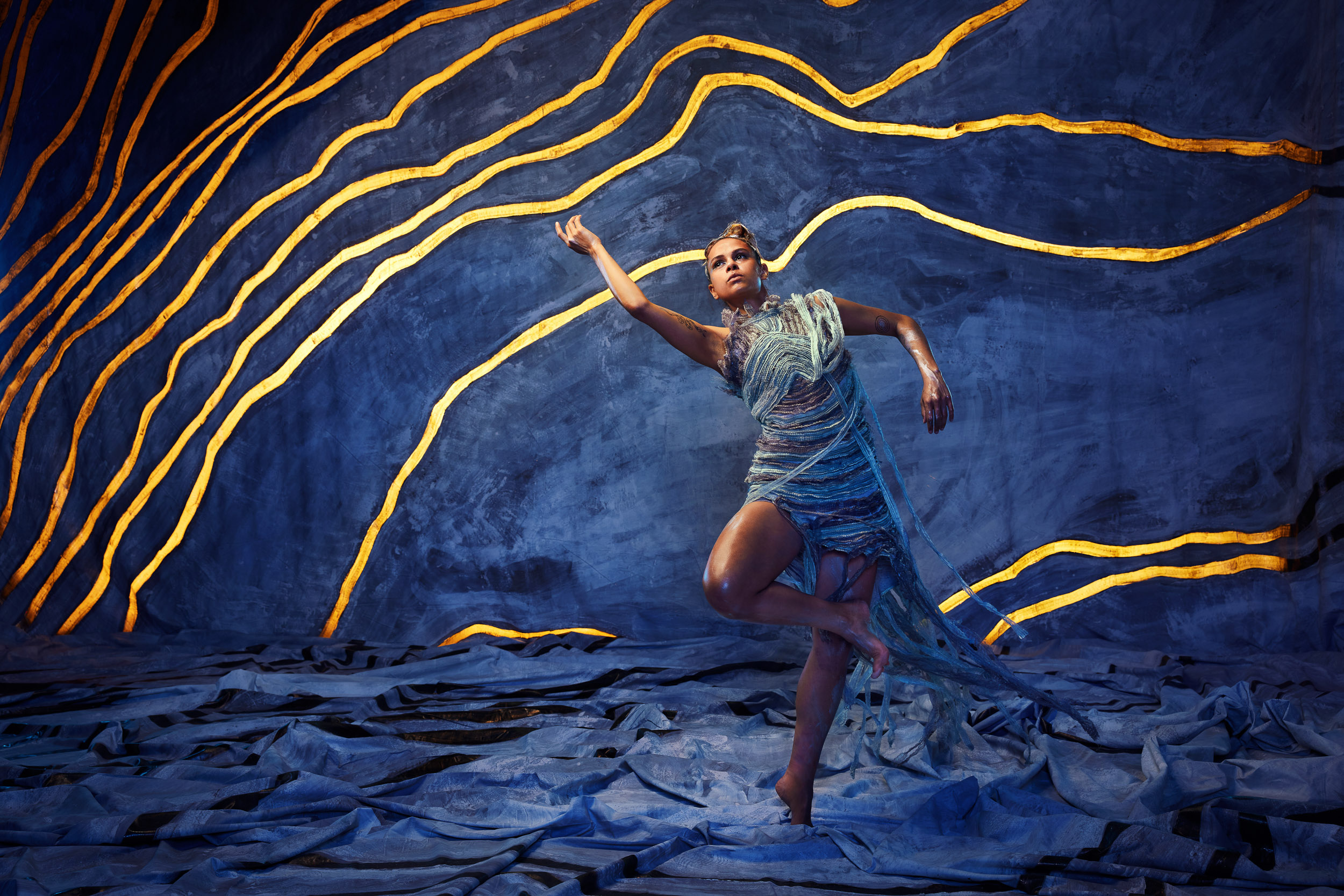 Elegant woman in flowing blue dress strikes a dynamic pose against an abstract blue and gold backdrop in a studio setting.
