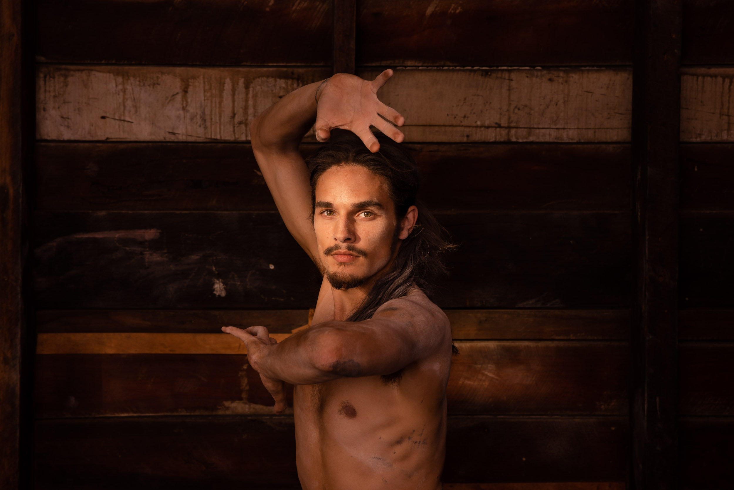 Adult male dancer poses with arms raised in a dimly lit indoor space with a rustic wooden background; dramatic lighting highlights his form.
