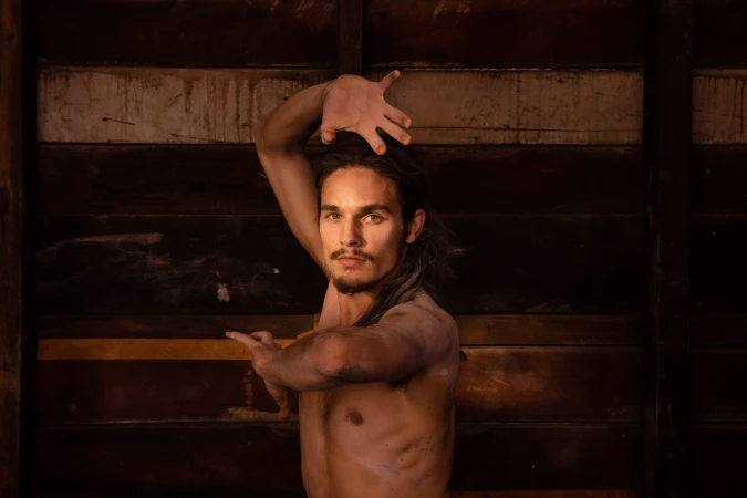 Adult male dancer poses with arms raised in a dimly lit indoor space with a rustic wooden background; dramatic lighting highlights his form.