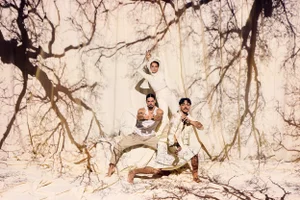 Three dancers posing against a cream backdrop with stark tree branch shadow projections, photographed by Daniel Boud for Bangarra Dance Theatre.