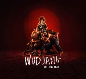 Group of dancers posed in a pyramid structure on cracked red earth with a large circular red spotlight behind them