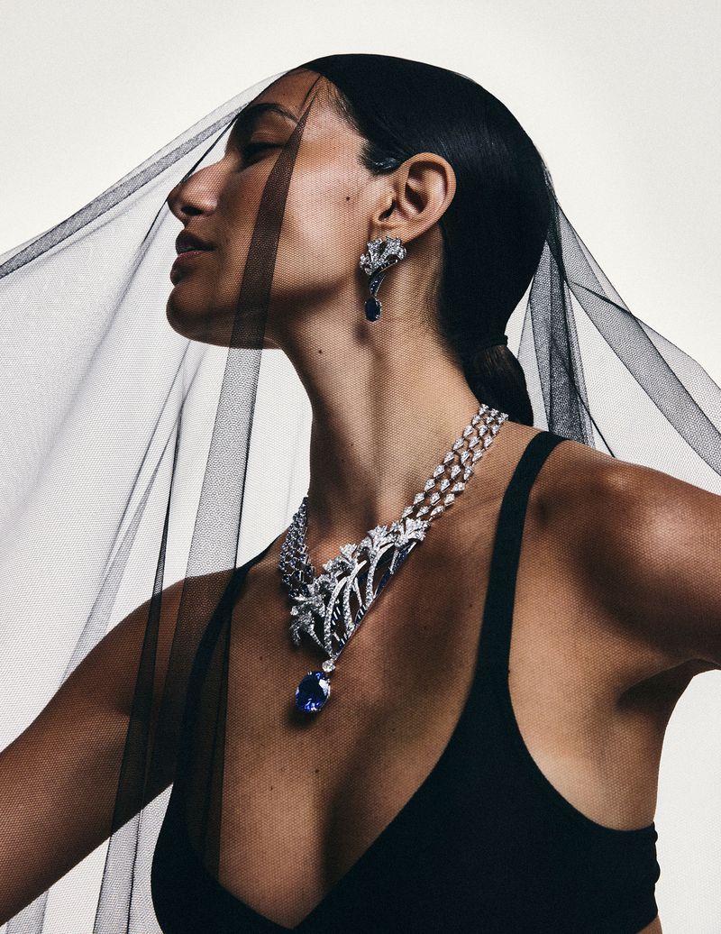 Woman with black hair poses in a black top with a large gem necklace and earrings, partially covered by a black veil.