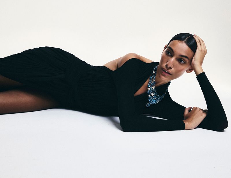 Fashion shot: an adult female model lies in a black dress with a star-themed necklace, shot in a photo studio with a white background.