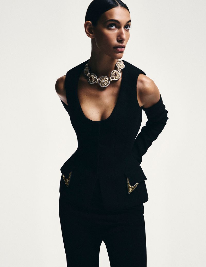 Woman in black suit with ornate gold necklace, wearing long gloves, posing in studio lighting. Fashion editorial, luxury aesthetic.