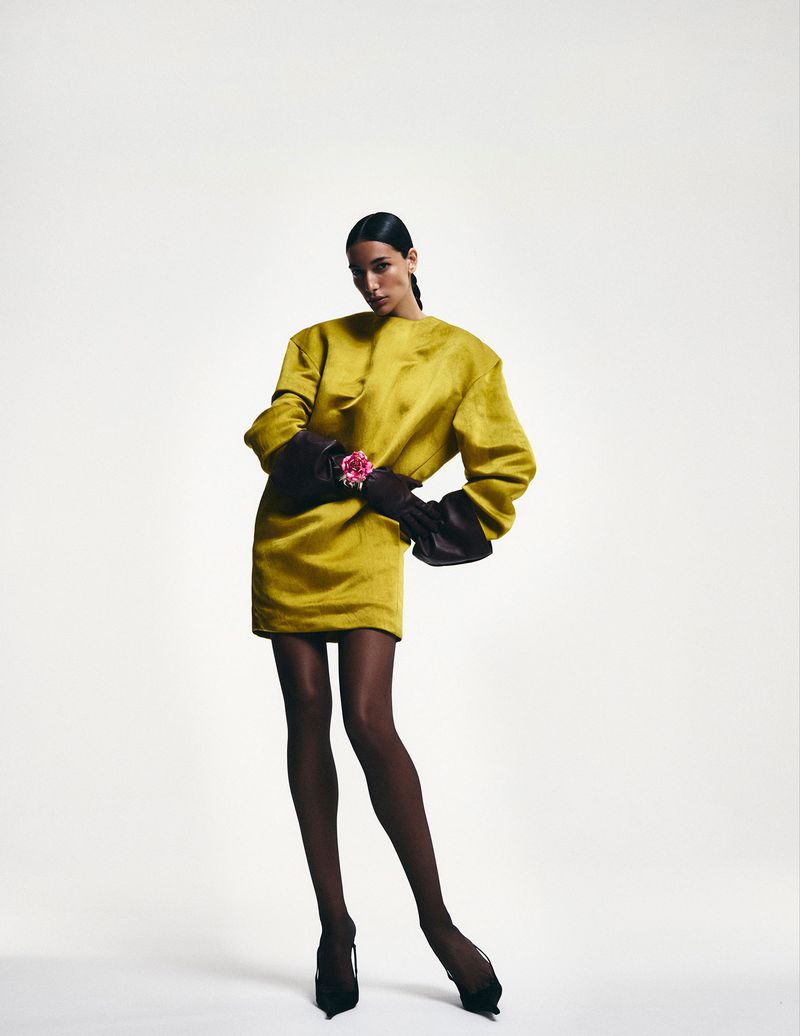 An adult female fashion model poses in a short gold dress with dark hosiery and heels against a white backdrop in a studio shot.