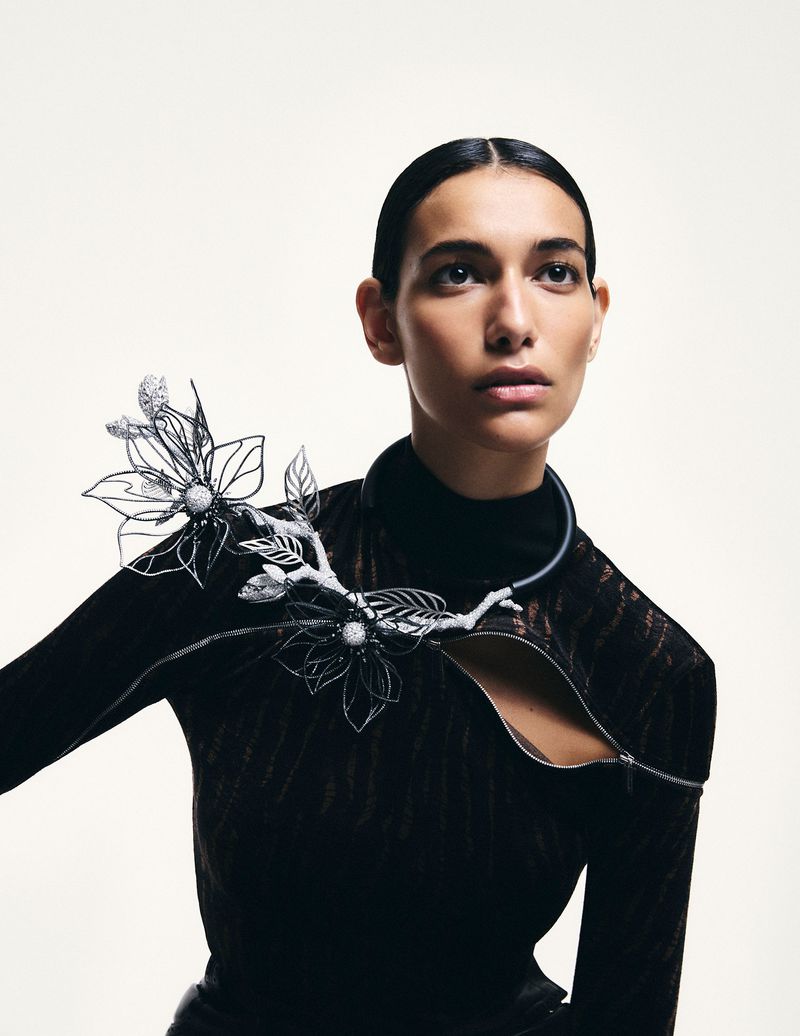 Close-up of a woman with slicked-back hair wearing a black top and a floral, branch-like necklace accessory against a white background.