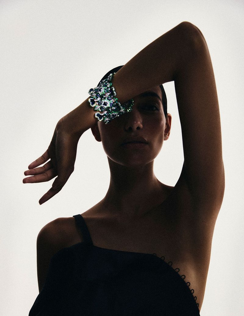 A woman in black dress covers her eyes with her hand and a sequined bracelet against a light background in a studio shot.