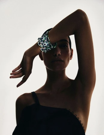 A woman in black dress covers her eyes with her hand and a sequined bracelet against a light background in a studio shot.