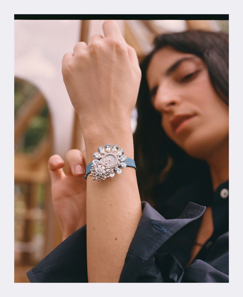 Woman's wrist wearing a designer watch with jeweled detailing, styled with a navy-blue button-up shirt in an outdoor setting.