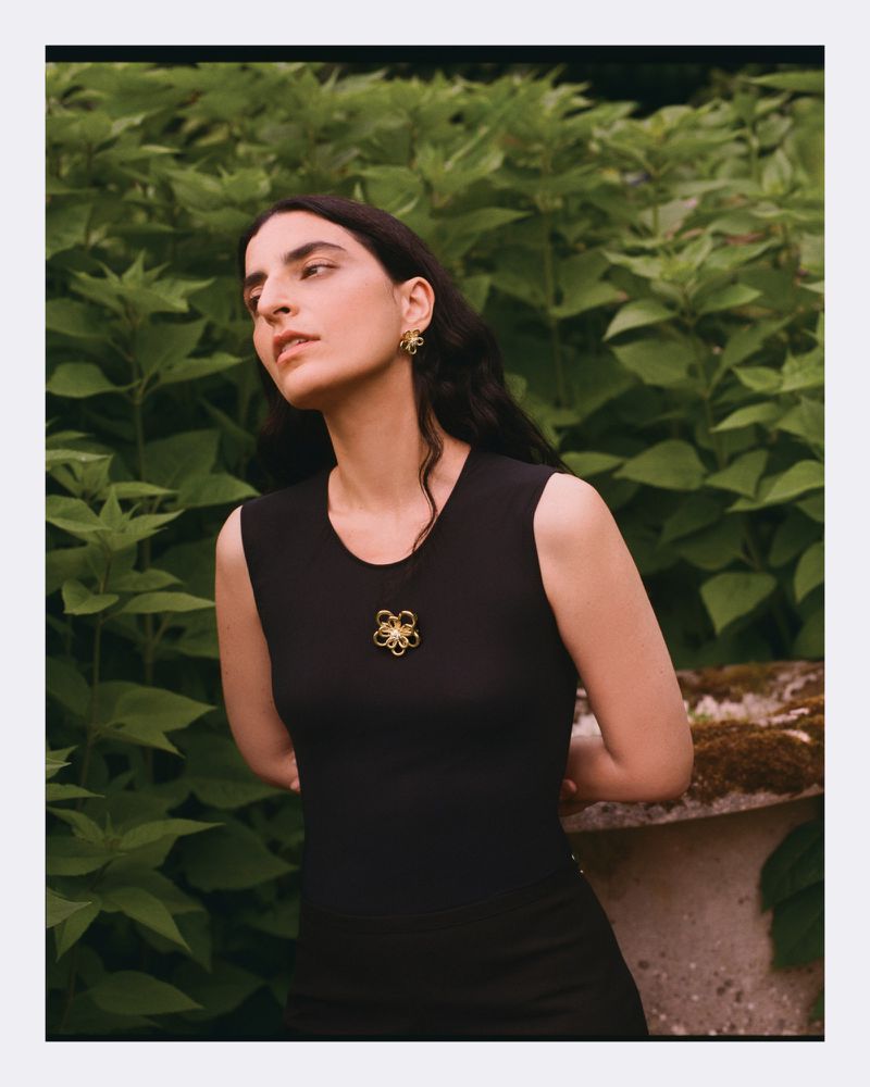 Elegant woman in a black dress poses in front of a lush green background, showcasing gold flower-shaped earrings and a matching necklace.