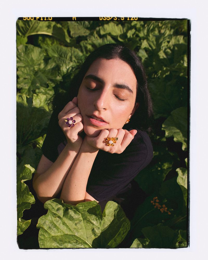 Woman with dark hair in a black shirt surrounded by large green leaves, showcasing rings with amber and amethyst stones.