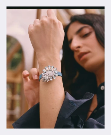 Close-up of a model's wrist wearing an ornate floral diamond watch with a blue strap against a softly blurred background.