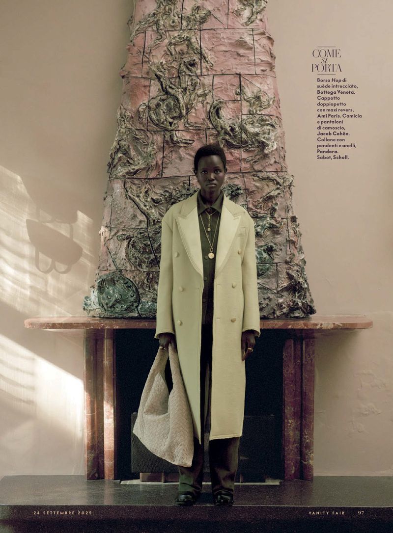 Fashion editorial shot of a Black woman posing indoors wearing a light-yellow coat and holding a beige handbag in muted lighting.