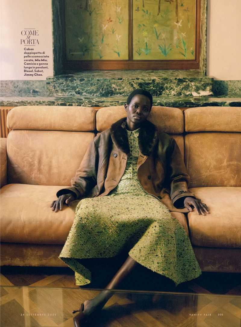 Fashion editorial shot of a Black woman wearing a green dress and brown jacket, sitting on an orange couch in a room with art.