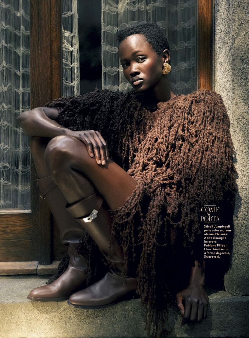 Fashion shot of a Black woman in a fringed shawl and leather boots sitting on a stone step, with a window behind her.