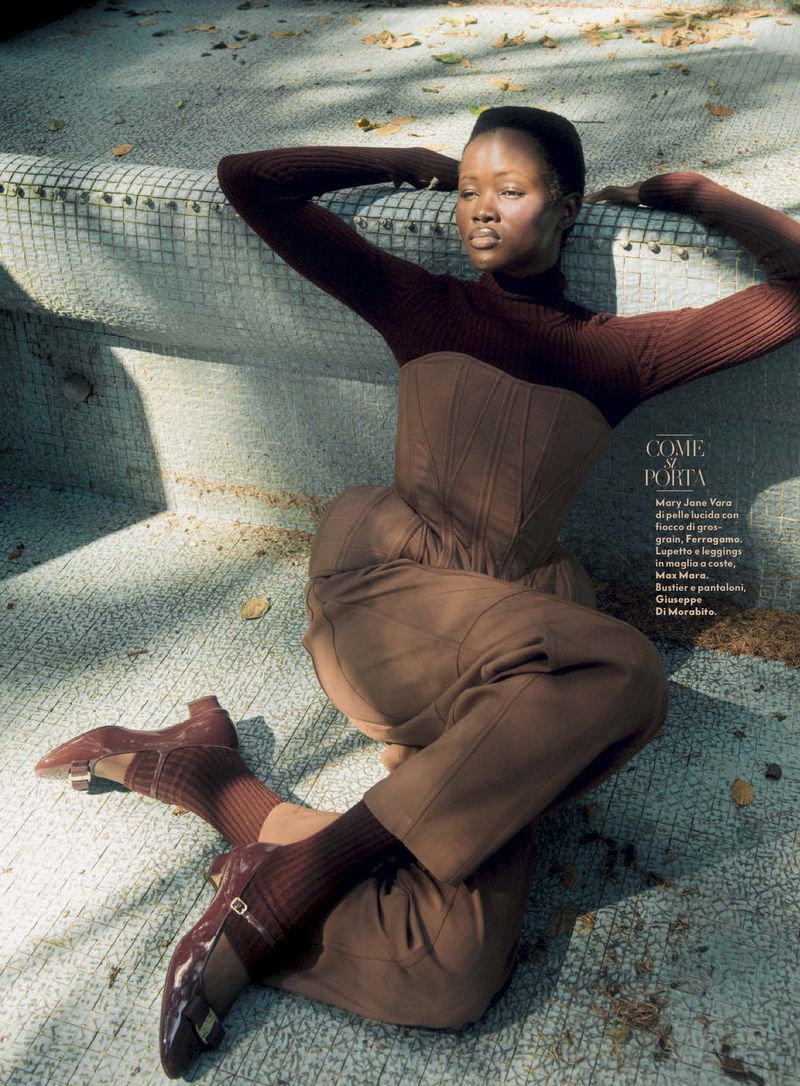A statuesque Black woman in a corset and matching trousers reclines in a geometric, tiled pool or fountain area outdoors on a sunny day.