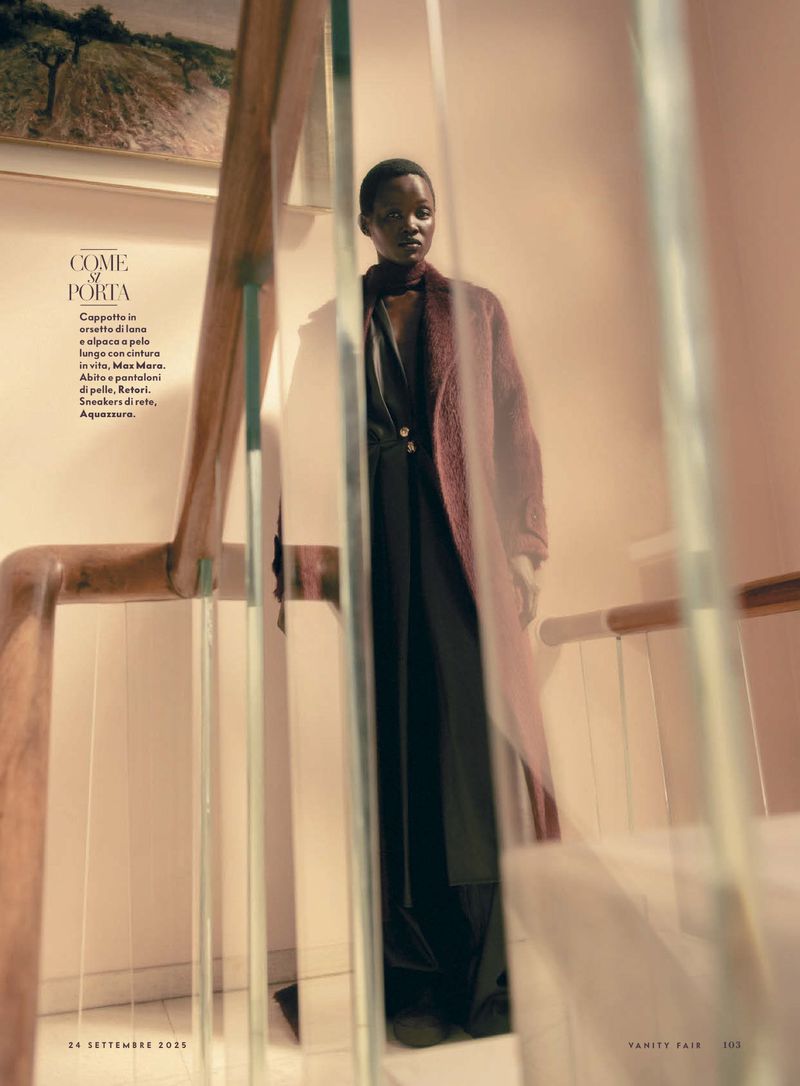 A fashion shot features a Black woman in a coat standing on a staircase inside, lit by natural light, creating a moody, elegant atmosphere.