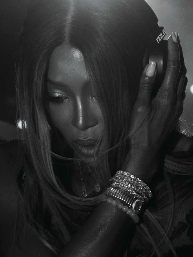 Black and white close up portrait of Naomi Campbell holding DJ headphones to her ear wearing diamond bracelets.