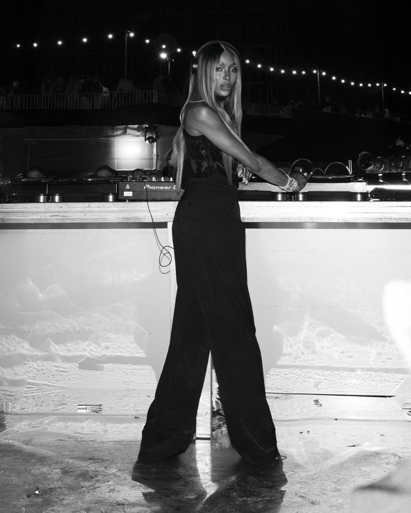 Black and white portrait of a woman in a dark outfit standing in front of DJ equipment with a crowd and string lights in the background.