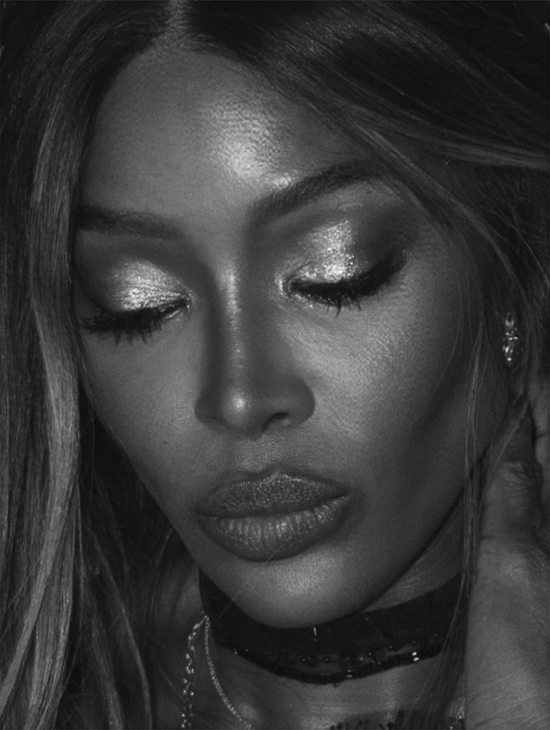 Black and white portrait of a woman with long hair, dramatic eye makeup, a choker necklace, and a chain necklace, looking downward.