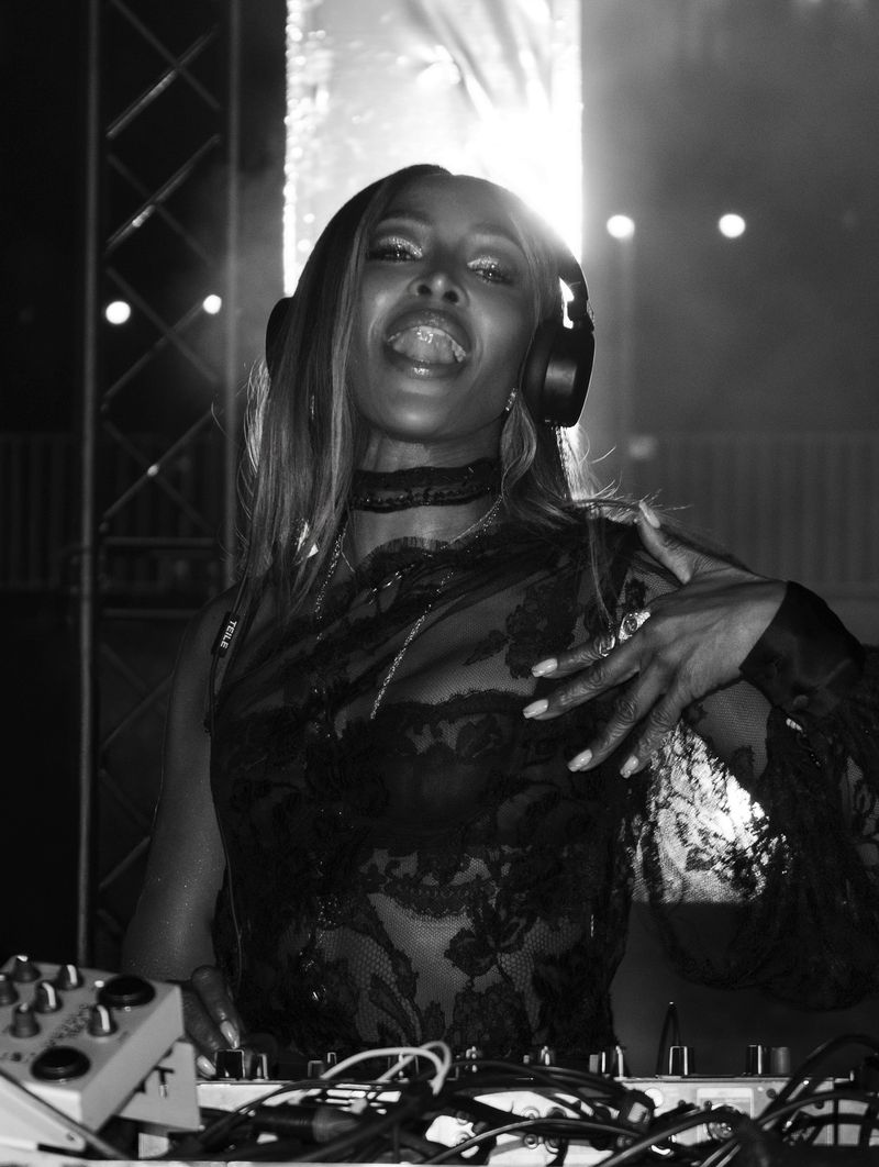Black and white portrait of an adult woman with long hair, wearing headphones and a sheer top, standing behind a mixing console.