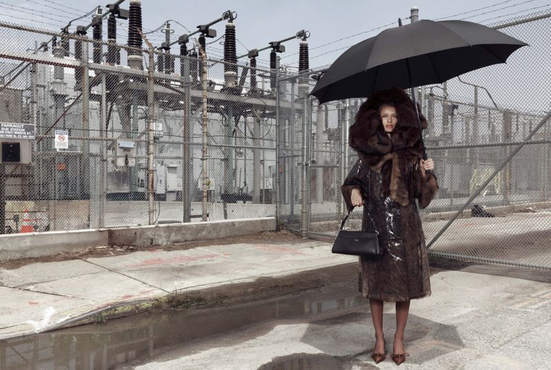 A woman in a faux fur-trimmed coat and umbrella stands on a city street near industrial electrical equipment, wet from recent rain.