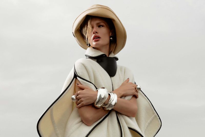 A woman in a cream cape and wide-brimmed hat poses against a soft grey sky, showcasing oversized silver bracelets and rings.