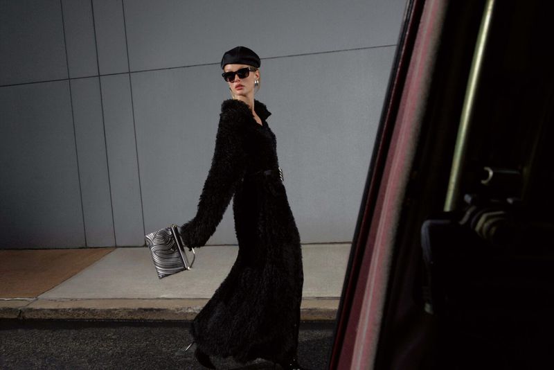 A stylish adult female in a long black coat, hat, and sunglasses poses with a patterned purse against a modern, urban backdrop.