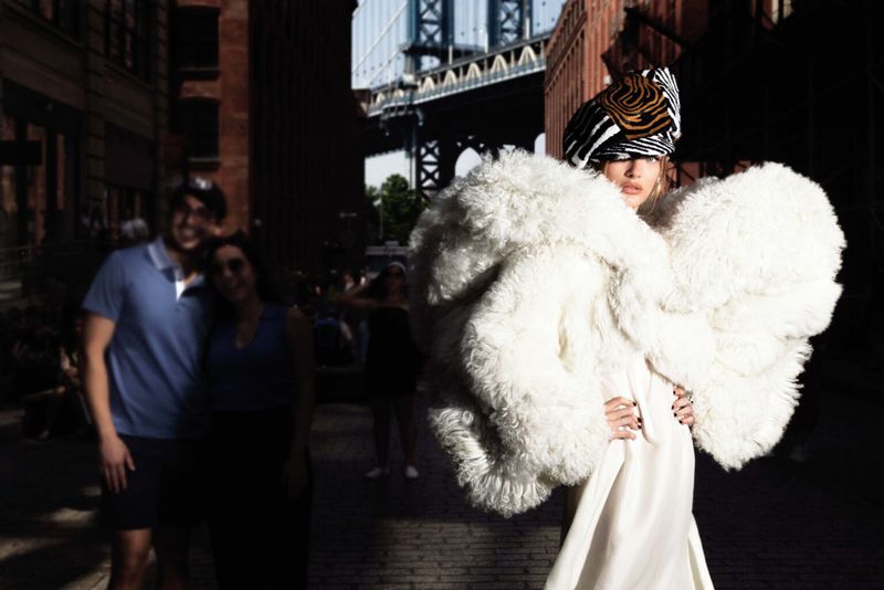 Fashion model in a fluffy white coat and patterned head wrap poses in an urban setting with a bridge in the background.