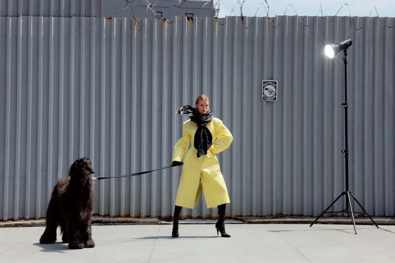 Outdoor fashion shoot featuring a model in a yellow coat with a dog, standing against a corrugated metal fence with studio lighting.