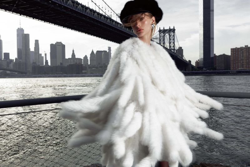 Fashionable woman in white faux fur coat poses against the backdrop of the Manhattan Bridge and the New York City skyline over water.