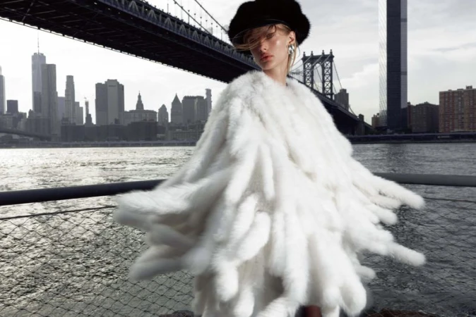 Fashionable woman in white faux fur coat poses against the backdrop of the Manhattan Bridge and the New York City skyline over water.