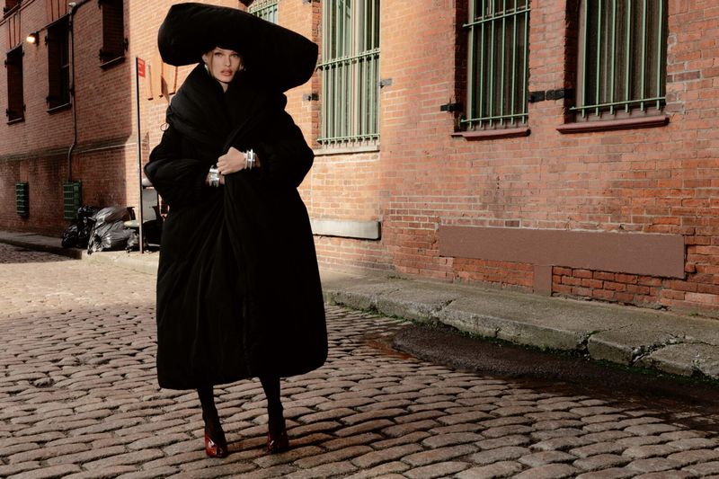 Woman poses in oversized black coat and hat on cobblestone street in urban alleyway. Fashion editorial showcases oversized style and texture.