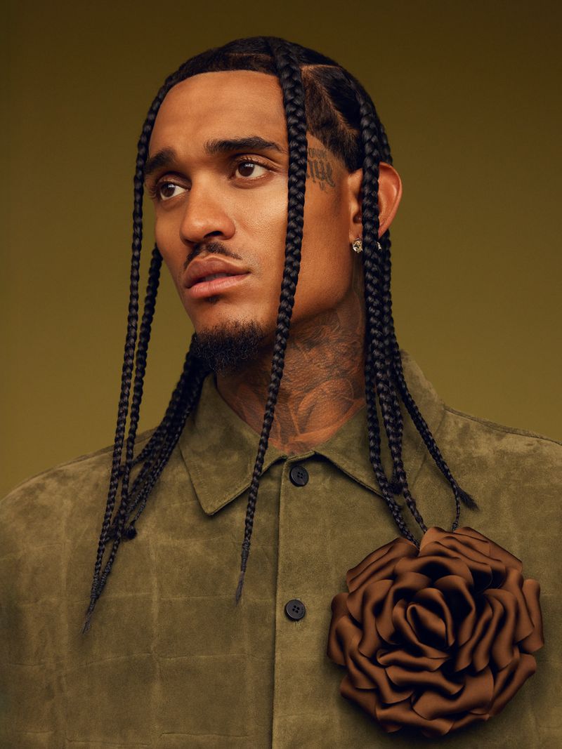 Portrait of an adult male with braided hair, wearing an olive green shirt and rose embellishment, looking to the side against a green background.