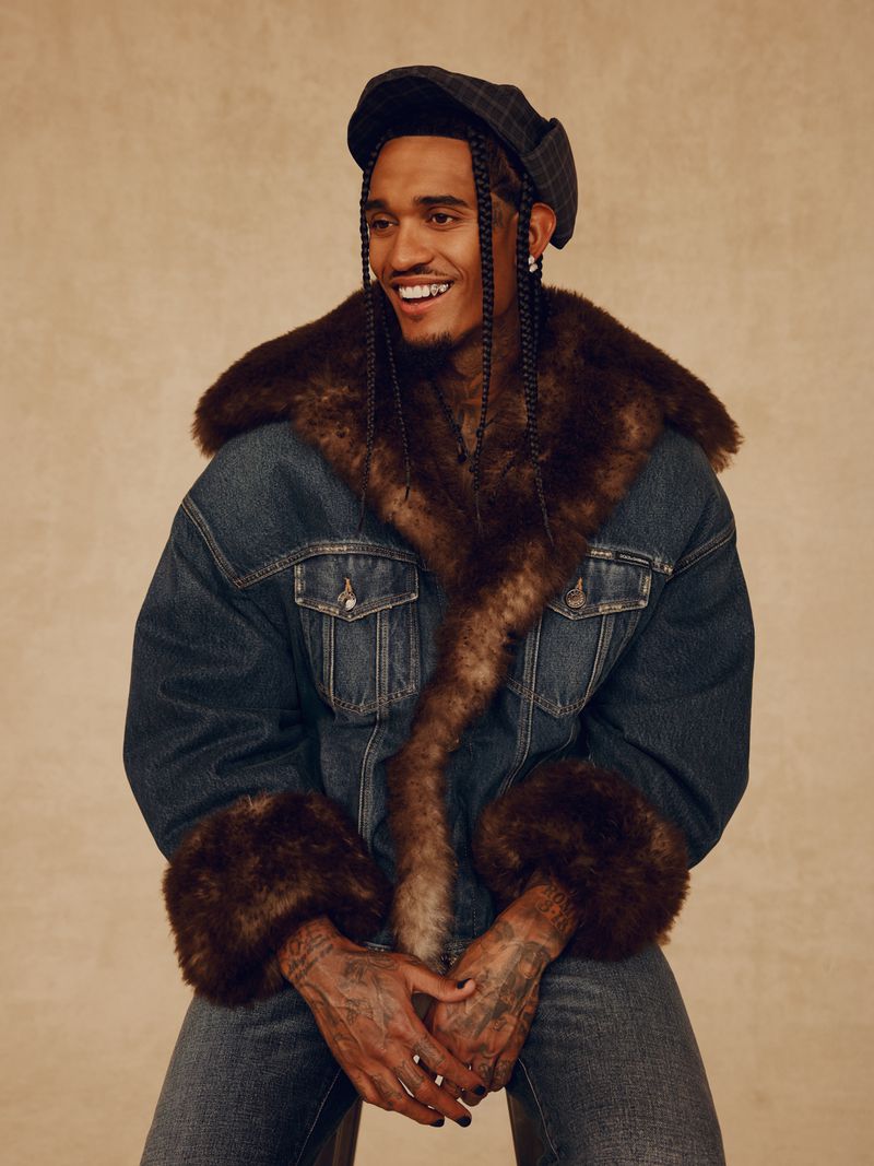 Portrait of a smiling adult male in a denim and fur jacket and newsboy cap sitting in a studio with a neutral background.