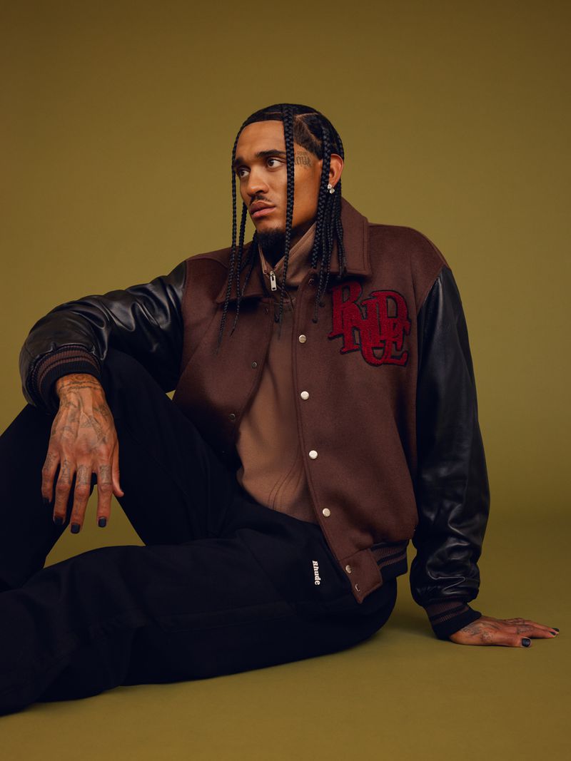 A man with braided hair sits in a brown jacket and black sweatpants against a muted green backdrop in a studio fashion shot.