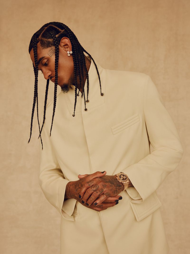 Portrait of an adult male with braids wearing an off-white suit and a gold watch, set against a neutral beige background.