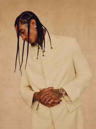 Portrait of an adult male with braids wearing an off-white suit and a gold watch, set against a neutral beige background.