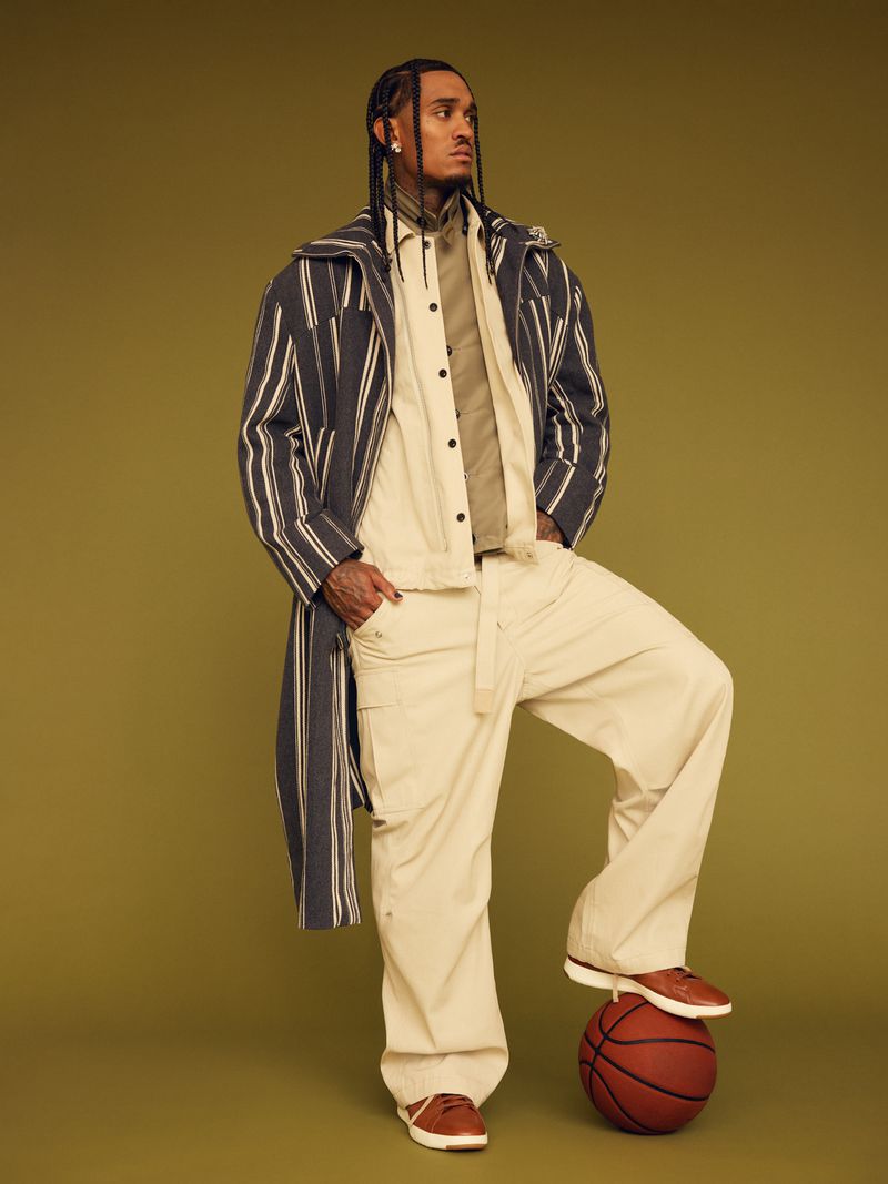 Man in fashionable striped overcoat, tan pants, brown shoes posing with foot on basketball in a studio with tan backdrop.